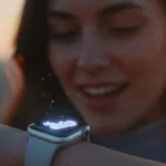 A woman with a soft, joyful expression looks down at her Apple Watch in the warm glow of sunrise. The watch on her wrist is illuminated, displaying a small, glowing holographic icon of a baby, creating an intimate and emotional scene.